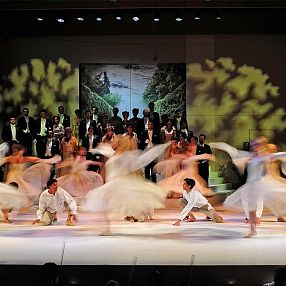 Bühnenauftritt mit Tänzern in weißen Kostümen, die sich in dynamischen Posen bewegen. Im Hintergrund steht ein Chor, beleuchtet von bunten Bühnenlichtern.