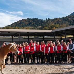 Eine Gruppe von Menschen steht in einer Reithalle vor einer bergigen Landschaft. Zwei Pferde laufen im Vordergrund. Alle tragen rot-schwarze Kleidung.
