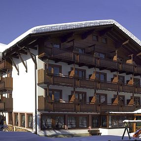 Großes, alpines Gebäude mit Holzelementen und Balkonen, umgeben von Schnee. Ein Spielplatz mit Rutsche ist im Vordergrund sichtbar. Klarer blauer Himmel.
