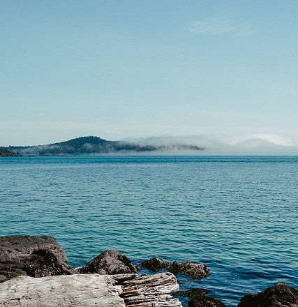 Ein weiter Blick über ein ruhiges Meer mit blauem Himmel. Im Hintergrund sind neblige Berge, teilweise in Wolken gehüllt, zu sehen. Natur und Ruhe dominieren.