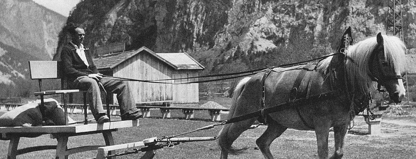 Historische schwarz-weiße Aufnahme zeigt ein Pferd, das einen Karren zieht. Im Hintergrund sind ein einfaches Holzgebäude und Felswände zu sehen.