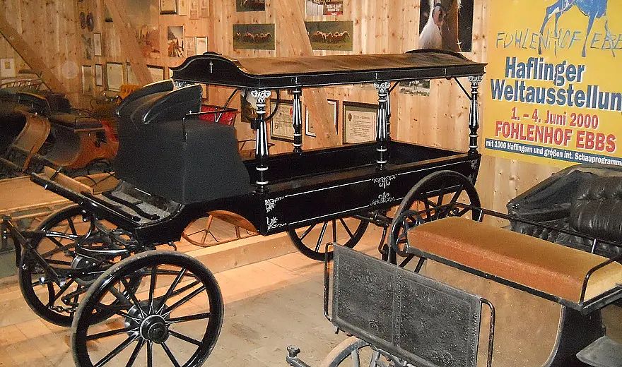 Historischer, schwarzer Pferdewagen im Holzmuseum. Nebenwägen und ein Poster zur Haflinger-Weltausstellung rahmen die Szene ein. Tradition trifft Geschichte.