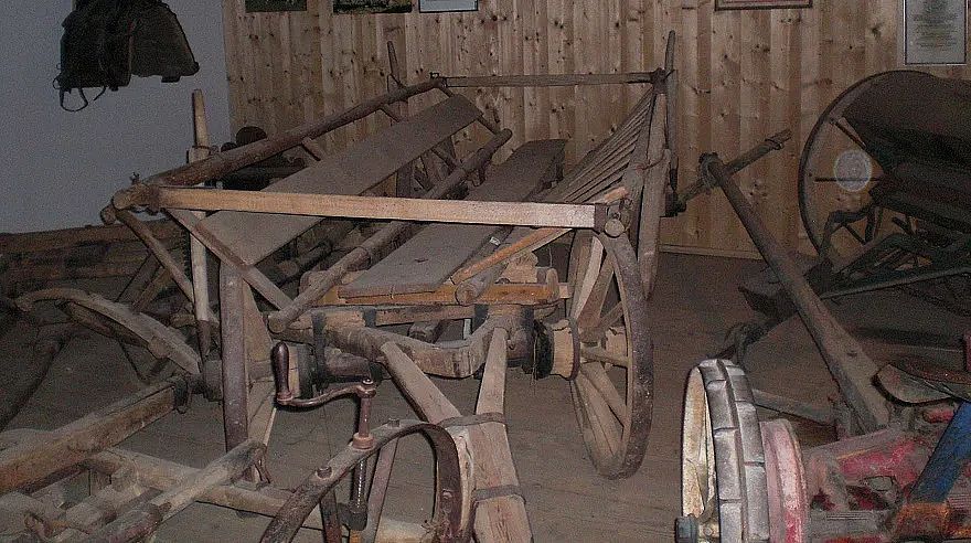 Altes landwirtschaftliches Gerät aus Holz mit zwei großen Rädern, vermutlich ein Heuwagen oder Pflug, in einem rustikalen Raum mit Holzwänden ausgestellt.