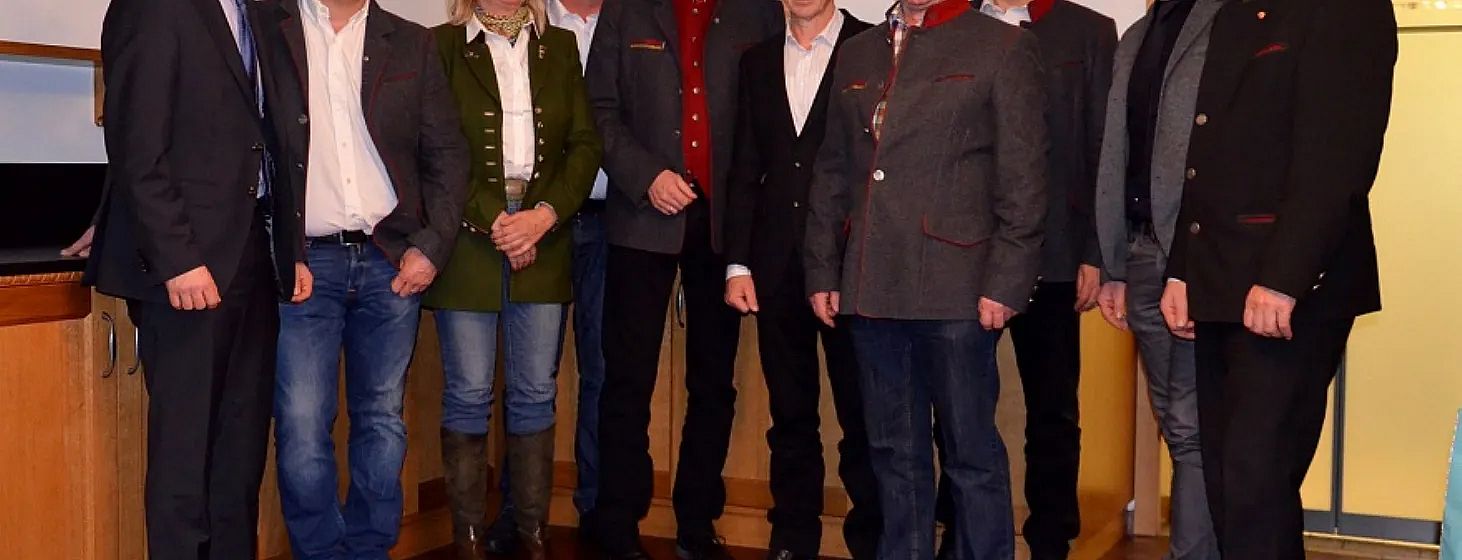 A group of people standing together, dressed in a mix of traditional and formal clothing, posing for a photo indoors against a plain background.