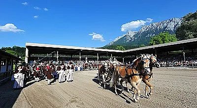 Pferdekutsche zieht festlich gekleidete Menschen auf einem weitläufigen Platz vor Bergkulisse. Zuschauer sind unter einem Vordach versammelt, blauer Himmel.