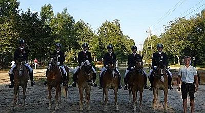 Sechs Reiter in Turnierkleidung sitzen auf Pferden, aufgereiht in einer Reihe. Im Hintergrund sind Bäume und ein klarer Himmel zu sehen.
