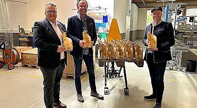 Drei Männer stehen in einer Werkstatt und halten goldene Hasenfiguren. Ein Tisch mit mehreren ähnlichen Figuren steht im Hintergrund. Alle lächeln freundlich.