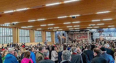 Großer Veranstaltungssaal mit Holzdecke, gefüllt mit vielen Menschen. Eine Bühne im Hintergrund mit der Aufschrift "Tegernsee" und Musikern. Teil einer Messe.