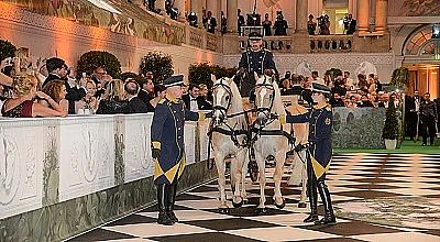 Elegante Veranstaltung mit zwei weißen Pferden, geführt von uniformierten Personen. Publikum in festlicher Kleidung beobachtet in opulentem, dekorierten Saal.