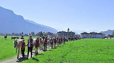 Menschen führen Pferde durch eine grüne Wiese vor einer Bergkulisse. Im Hintergrund sind Gebäude und ein Kirchturm zu sehen, der Himmel ist klar.