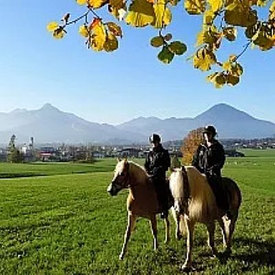 Zwei Reiter auf braunen Pferden, umgeben von herbstlichen Bäumen und grünen Feldern, mit Bergen im Hintergrund unter einem klaren blauen Himmel.