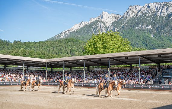 Eine Gruppe von Haflingerpferden führt eine Show in einer offenen Arena vor malerischer Bergkulisse auf. Ein Publikum sitzt auf überdachten Tribünen und schaut zu.