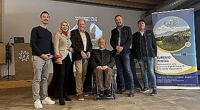 Sechs Personen stehen nebeneinander bei einer Veranstaltung. Ein Mann sitzt im Rollstuhl. Im Hintergrund sind eine Leinwand und ein Banner zu sehen.