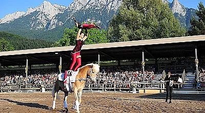 Eine Akrobatin balanciert auf einem Pferd in einer Arena vor einem Publikum, mit malerischen Bergen und grünen Bäumen im Hintergrund.