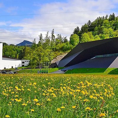 Modernes Gebäude mit geometrischen Formen vor einer malerischen Berglandschaft und blühender Wiese. Das weiße und schwarze Design kontrastiert die Natur.